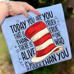 May include: Light blue t-shirt featuring a whimsical illustration of a red and white striped hat. The shirt has the text "TODAY YOU ARE YOU TRUER THAN THERE IS ALIVE IS YOUER THAN YOU" in black, stylized lettering.