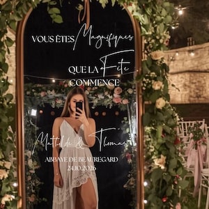 Peut inclure: Un grand miroir en forme d'arche avec un cadre doré est orné de fleurs et de verdure. Le miroir reflète une personne en robe blanche. Le texte sur le miroir dit "Vous êtes Magnifiques, Que la Fête Commence" et des noms et une date.