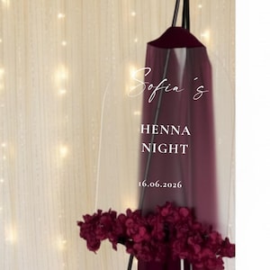 Op de afbeelding: Een bordeauxrood Henna Night-bord met de naam "Sofia's" in elegant schrift. Het bord is versierd met een tros bordeauxrode bloemen aan de basis en staat tegen een achtergrond van een crèmekleurig gordijn met feeërieke lichtjes.