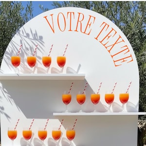 Op de afbeelding: Een witte boogvormige displaystandaard met vier horizontale planken met negentien heldere glazen gevuld met een oranje vloeistof en rood-wit gestreepte rietjes. De tekst VOTRE TEXTE is in het oranje gedrukt bovenaan het witte paneel.
