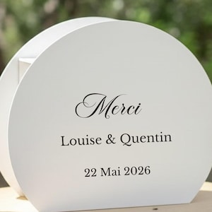 Könnte beinhalten: Eine weiße, kreisförmige Holzbox mit dem schwarzen Aufdruck "Merci", "Louise & Quentin" und "22 Mai 2026" auf der Vorderseite. Das Objekt hat eine glatte, matte Oberfläche und eine geschwungene, halbzylindrische Form.