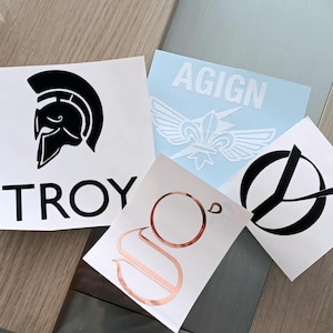 Könnte beinhalten: Eine Sammlung von Vinyl-Aufklebern mit verschiedenen Designs. Ein Aufkleber zeigt einen schwarzen spartanischen Helm mit dem Wort "TROY" darunter. Weitere Aufkleber zeigen ein weißes Design auf hellblauem Hintergrund, ein roségoldenes Design und ein schwarz-weißes Design.
