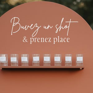 May include: A terracotta-colored shot glass display with the text "Buvez un shot & prenez place" in white script. The display has three shelves holding clear shot glasses, perfect for a wedding or event.