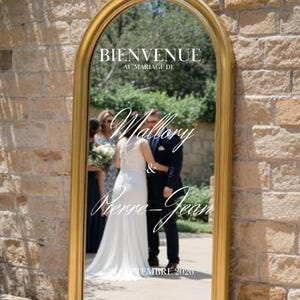 以下が含まれることがあります： 上部に「BIENVENUE AU MARIAGE DE」の文字が入った、大きなアーチ型の金色のフレームの鏡。鏡には花嫁と花婿が映っており、「Mallory & Pierre-Jean」という名前と「SEPTEMBRE 2020」という日付が書かれています。