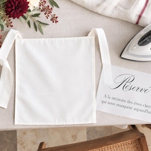 Peut inclure: Un panneau de tissu blanc carré avec deux longs liens en tissu aux coins supérieurs. À côté se trouve une carte blanche avec le texte imprimé en noir : Réservé, A la mémoire des êtres chers qui nous manquent aujourd'hui, et un fer à repasser.