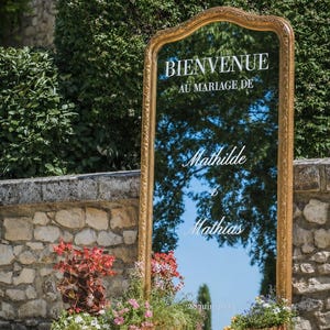 Peut inclure: Un grand miroir rectangulaire sur pied avec un cadre doré orné. La surface en verre porte l'inscription blanche BIENVENUE AU MARIAGE DE Mathilde & Mathias 28 juin 2024. Le miroir reflète le ciel et est entouré de plantes en pot.