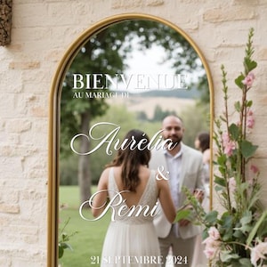 Puede incluir: Espejo con marco dorado con las palabras "BIENVENUE AU MARIAGE DE Aurélia & Rémi 21 SEPTEMBRE 2024" en escritura blanca. El espejo refleja a una novia y un novio, con arreglos florales a la derecha.