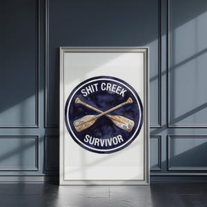 Könnte beinhalten: Gerahmter Kunstdruck mit dem Text "SHIT CREEK SURVIVOR" und gekreuzten Rudern in einem marineblauen Kreis. Das Kunstwerk befindet sich in einem silbernen Rahmen vor einer dunkelblauen Wand und einem Boden.