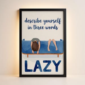 May include: A framed art print with a watercolor illustration. The image features a person lying on a blue couch with the word "LAZY" below. The text at the top reads "describe yourself in three words". The frame is black.