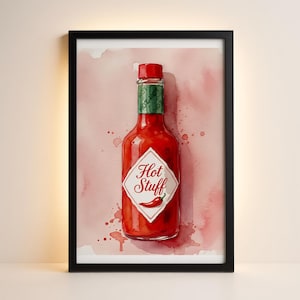 May include: A framed watercolor print of a red hot sauce bottle with a red cap and a green label. The label reads "Hot Stuff" with a chili pepper illustration. The background is a watercolor wash of pink and red.