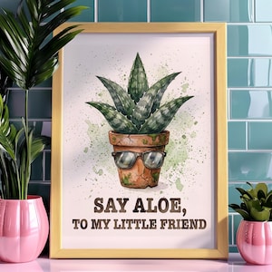 Puede incluir: Una impresión artística enmarcada con una ilustración de acuarela de una planta de aloe vera en una maceta de terracota con gafas de sol. El texto dice "Say Aloe, To My Little Friend". El marco es dorado.