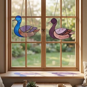 Pode incluir: Dois ornamentos de pato em vitral pendurados em frente a uma janela. Um pato é azul e roxo, o outro é roxo e rosa. Os patos são suspensos por correntes. A janela é de madeira e o fundo é uma cena externa desfocada.