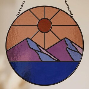 Pode incluir: Painel redondo de vitral retratando uma paisagem montanhosa ao pôr do sol. O design apresenta uma moldura castanha escura, um sol com raios, montanhas roxas e um lago azul. Suspenso por uma corrente, cerca de 20 cm de diâmetro.