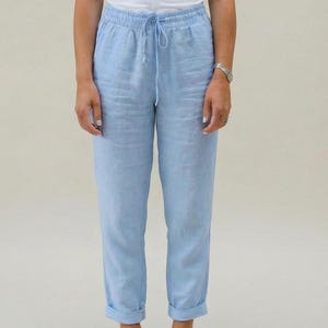 May include: A woman wearing a white tank top and light blue linen pants with a drawstring waist. The pants are cuffed at the ankles. The background is a solid, neutral color.