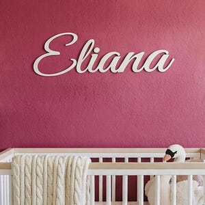 Könnte beinhalten: Ein weißes Wand-Namensschild aus Holz mit dem Namen Eliana in Schreibschrift. Das Schild ist an einer dunkelrosa Wand über einem Kinderbett aus hellem Holz und Weiß angebracht. Im Bett liegen eine weiße Strickdecke und ein Schwan aus Plüsch.