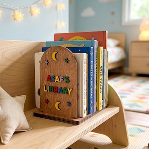 May include: A wooden bookend with an arched top, featuring the text ASAF'S LIBRARY in colorful letters, accented with cut-out stars and moons. The bookend holds a row of children's books on a light-colored wooden shelf next to a plush bear.