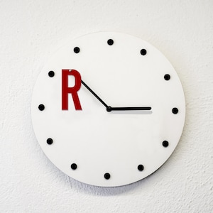 Può includere: Un orologio da parete bianco con indicatori delle ore a punti neri e lancette nere. La lettera "R" in rosso sostituisce il numero 1 sul quadrante. Le lancette indicano che sono circa le 3:15.