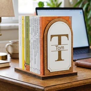 May include: A wooden bookend featuring an arched design with a white insert. The insert displays a large letter T with the name Tom printed horizontally across the center. The bookend supports a row of various books on a wooden desk surface.