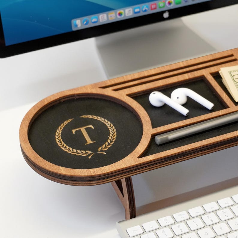 May include: A wooden desk organizer with a black surface and a light brown frame. The organizer has a circular section with a gold letter "T" inside a laurel wreath, and compartments for small items, including a pair of white earbuds and a stylus.