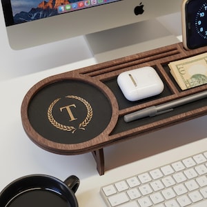 May include: A wooden desk organizer with multiple compartments. It features a round section with a gold-colored letter "T" inside a laurel wreath. Other sections hold a pen, money, and a pair of white wireless earbuds. A keyboard and computer monitor are in the background.