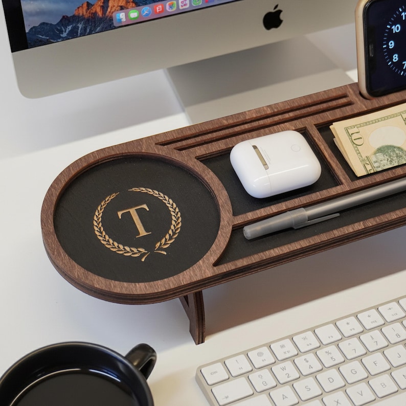 May include: A wooden desk organizer with multiple compartments. It features a round section with a gold-colored letter "T" inside a laurel wreath. Other sections hold a pen, money, and a pair of white wireless earbuds. A keyboard and computer monitor are in the background.