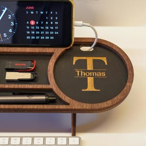 May include: A wooden desk organizer with a phone charging, a calendar display, and storage for small items. The organizer has a circular section with the name "Thomas" and the letter "T" engraved. A USB drive and pen are visible.