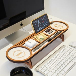 May include: A wooden desk organizer with multiple compartments, including a phone holder, pen slots, and a small tray. The organizer holds a smartphone displaying a clock, cash, pens, and other small items. The design features a light wood finish and a minimalist aesthetic.