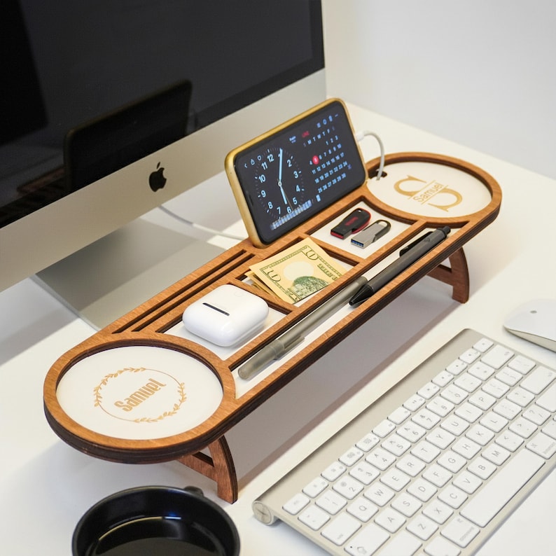 May include: A wooden desk organizer with multiple compartments, including a phone holder, pen slots, and a small tray. The organizer holds a smartphone displaying a clock, cash, pens, and other small items. The design features a light wood finish and a minimalist aesthetic.