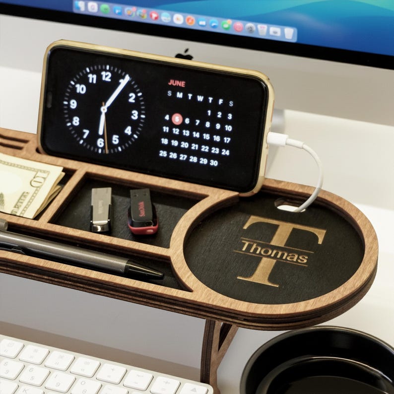 May include: Wooden desk organizer with a phone charging station. The phone displays a clock and calendar. Includes compartments for pens, USB drives, and a personalized name. The organizer is made of light brown wood.