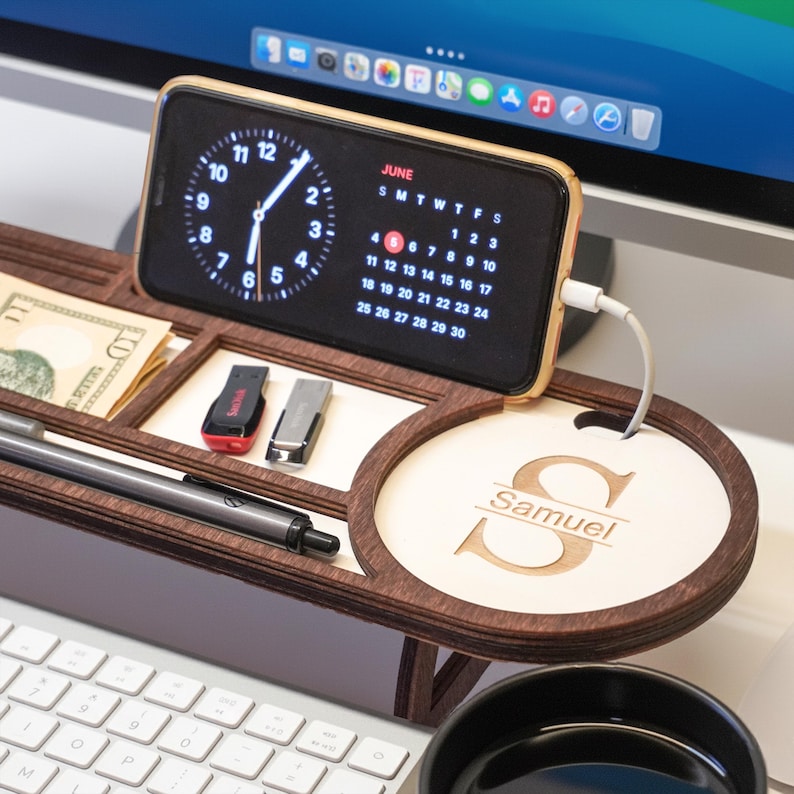 May include: A wooden desk organizer with a phone charging, a pen, cash, and USB drives. The phone displays a clock and calendar. The organizer has a personalized round section with the name "Samuel".