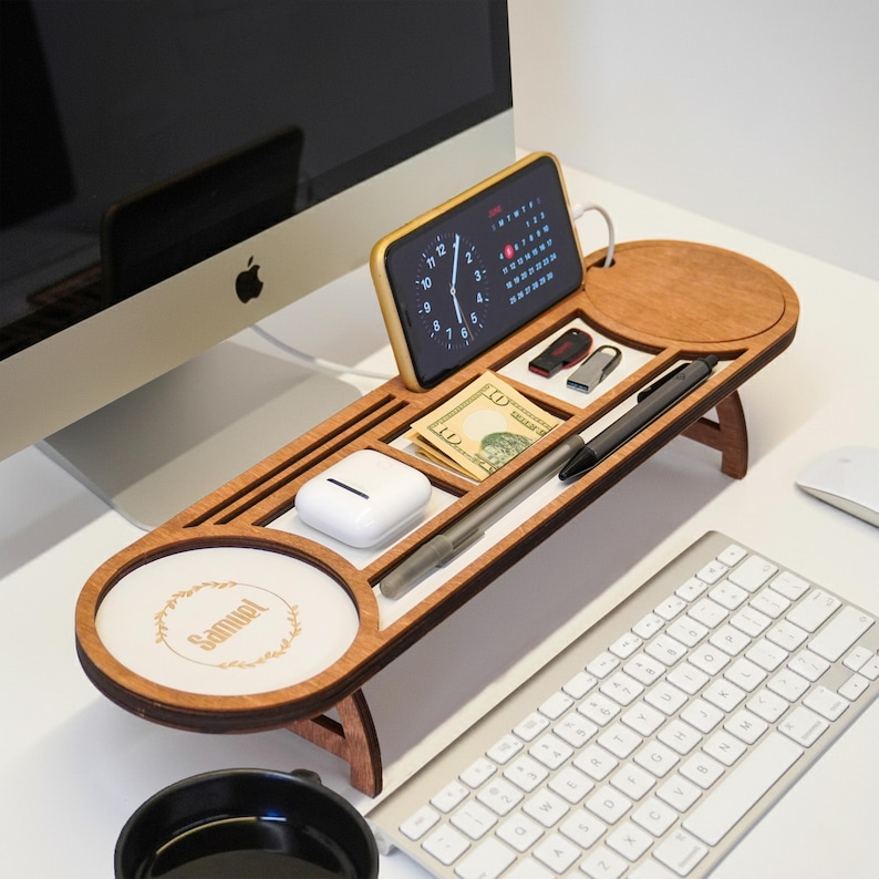 May include: A wooden desk organizer with multiple compartments, holding a smartphone, pens, cash, and other small items. The organizer has a circular section with the name "Samuel" and a floral design. The desk organizer is on a white desk.