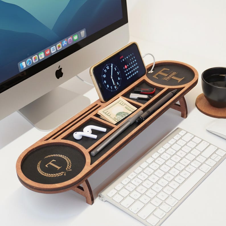 May include: A wooden desk organizer with multiple compartments, including a phone holder, pen slot, and small trays. The organizer features a black surface with gold-colored initials and a laurel wreath design. A smartphone displays a clock and calendar.