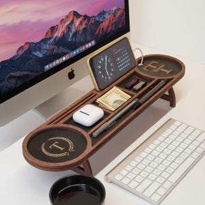 May include: A wooden desk organizer with multiple compartments for a phone, pens, cash, and other small items. The organizer has a dark wood finish with a black accent and a gold initial "T" design. A keyboard and computer monitor are in the background.