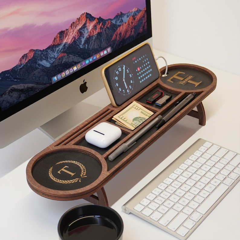 May include: A wooden desk organizer with multiple compartments for a phone, pens, cash, and other small items. The organizer has a dark wood finish with a black accent and a gold initial "T" design. A keyboard and computer monitor are in the background.