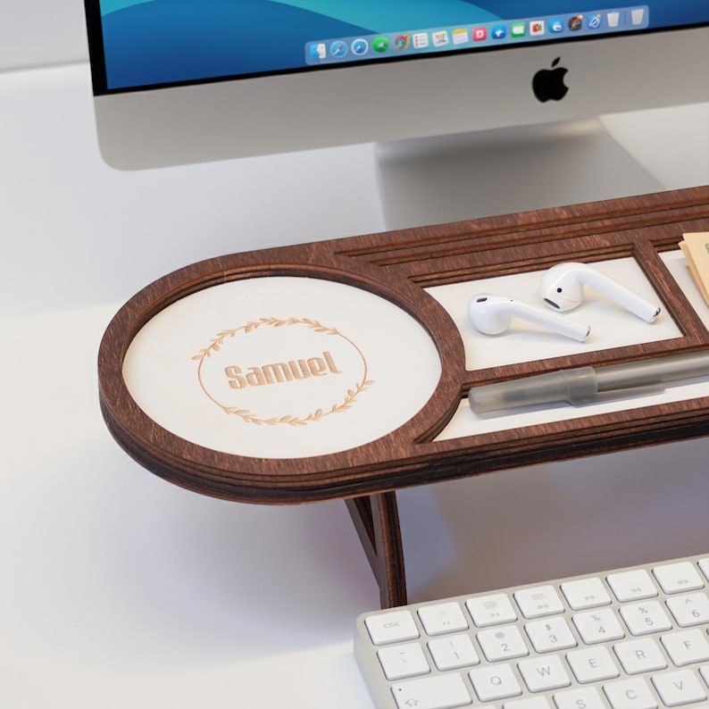 May include: A wooden desk organizer with a personalized circular section that reads "Samuel" surrounded by a leaf design. The organizer has a dark brown frame and a white surface, holding a pen and white earbuds. A computer keyboard is in the foreground.