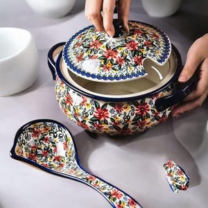 Może przedstawiać: Ceramiczna waza do zupy z pasującą chochlą i pokrywką, z kwiatowym wzorem w kolorze czerwonym, żółtym i zielonym na białym tle. Waza ma ciemnoniebieskie uchwyty i wykończenia. Pokrywka ma wycięcie na chochlę.