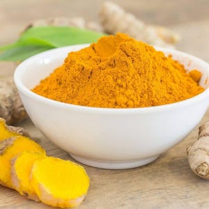 May include: A white ceramic bowl filled with a mound of bright orange-yellow turmeric powder. Surrounding the bowl are several raw, knobby turmeric roots, some of which are sliced into circular pieces, revealing a vibrant yellow interior.