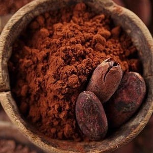Puede incluir: Primer plano de un cuenco de madera lleno de cacao en polvo marr&oacute;n oscuro y varios granos de cacao. El polvo tiene una textura fina y los granos son de color marr&oacute;n oscuro con una superficie texturizada. La imagen destaca los ingredientes del chocolate.