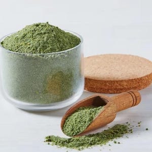 May include: A clear glass container filled with green powder, a wooden scoop with green powder, and a round, tan, porous object. The green powder is spilling out of the scoop and container. The objects are on a white surface.