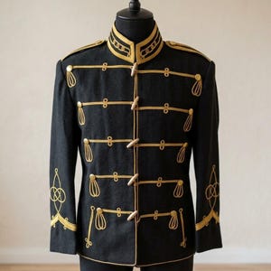 Könnte beinhalten: Schwarze Jacke im Militärstil mit goldenen Verzierungen und Akzenten. Die Jacke hat einen hohen Kragen, dekorative goldene Seilverschlüsse und goldene Stickereien an Schultern und Ärmeln. Die Jacke wird auf einer schwarzen Schaufensterpuppe präsentiert.