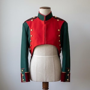 Op de afbeelding: Een militair-achtige jas in rood en groen, met gouden knopen en sierlijsten. De jas heeft een hoge kraag en lange mouwen. Het wordt getoond op een mannequin.
