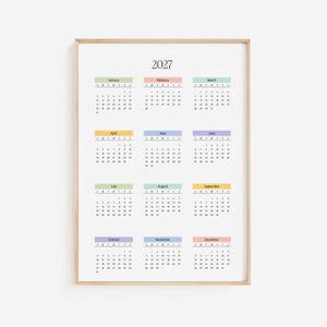 Peut inclure: Calendrier encadré de 2027 avec un cadre en bois clair. Chaque mois est affiché avec un en-tête de couleur pastel. La grille du calendrier est noire sur fond blanc. L'année "2027" est imprimée en gras noir en haut.
