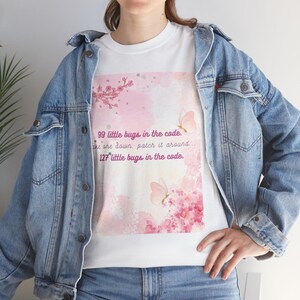 Peut inclure: T-shirt blanc avec un motif aquarelle rose représentant des papillons et des fleurs de cerisier. Le texte dit : "99 petits bogues dans le code. On en prend un, on le corrige... 127 petits bogues dans le code."