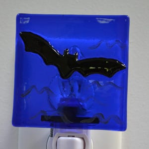 May include: A blue glass night light with a black bat design. The bat is centered on the blue glass, with wavy lines below. The night light is plugged into a white outlet.