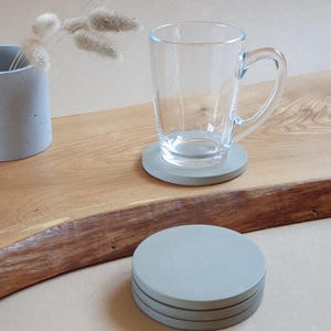 May include: A clear glass mug sits on a gray concrete coaster. A stack of three similar coasters is in the foreground. A small gray concrete vase with dried flowers is to the left. The items are on a wooden surface.