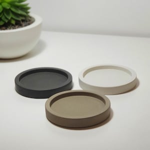 Könnte beinhalten: Drei runde Untersetzer in Schwarz, Weiß und Beige. Die Untersetzer bestehen aus einem festen Material und haben einen leicht erhöhten Rand. Ein weißer Keramiktopf mit einer grünen Sukkulentenpflanze befindet sich im Hintergrund.