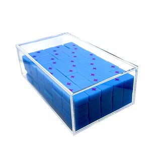 May include: A clear acrylic box filled with blue rectangular tiles. Each tile has a purple star design. The tiles are stacked neatly inside the box, creating a visually appealing arrangement. The box is on a white background.