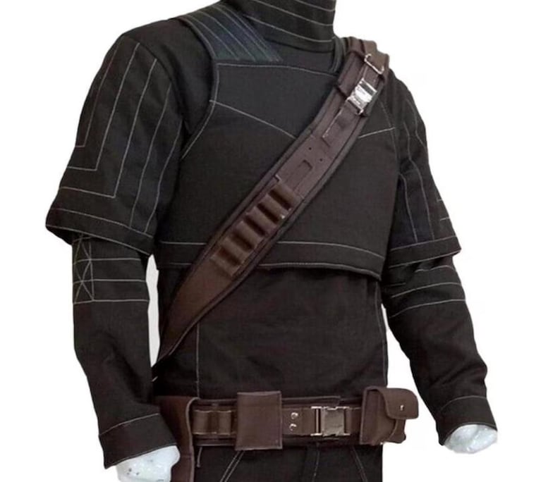 Op de afbeelding: Zwarte tactische jas met een hoge kraag, witte stiksels en een bruine leren schouderriem met munitiehouders. De jas heeft een bijpassende riem met zakjes en een zilveren gesp. De mouwen hebben een gelaagd ontwerp.