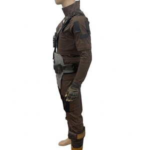 K&ouml;nnte beinhalten: Ein brauner taktischer Anzug mit hohem Kragen, langen &Auml;rmeln und mehreren Taschen. Das Outfit umfasst einen G&uuml;rtel, Handschuhe und Beinbedeckungen. Das Kost&uuml;m ist f&uuml;r eine robuste, zweckm&auml;&szlig;ige &Auml;sthetik konzipiert.