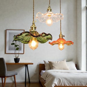 Könnte beinhalten: Drei Pendelleuchten mit blütenförmigen Glasschirmen in Grün, Klar und Orange. Jede Leuchte hat eine Glühbirne im Vintage-Stil und Messingbeschläge. Die Leuchten hängen an braunen Kabeln und verleihen dem Raum Eleganz.